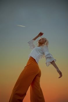 Stylish woman with arms raised, wearing orange pants and white top against a sunset sky.