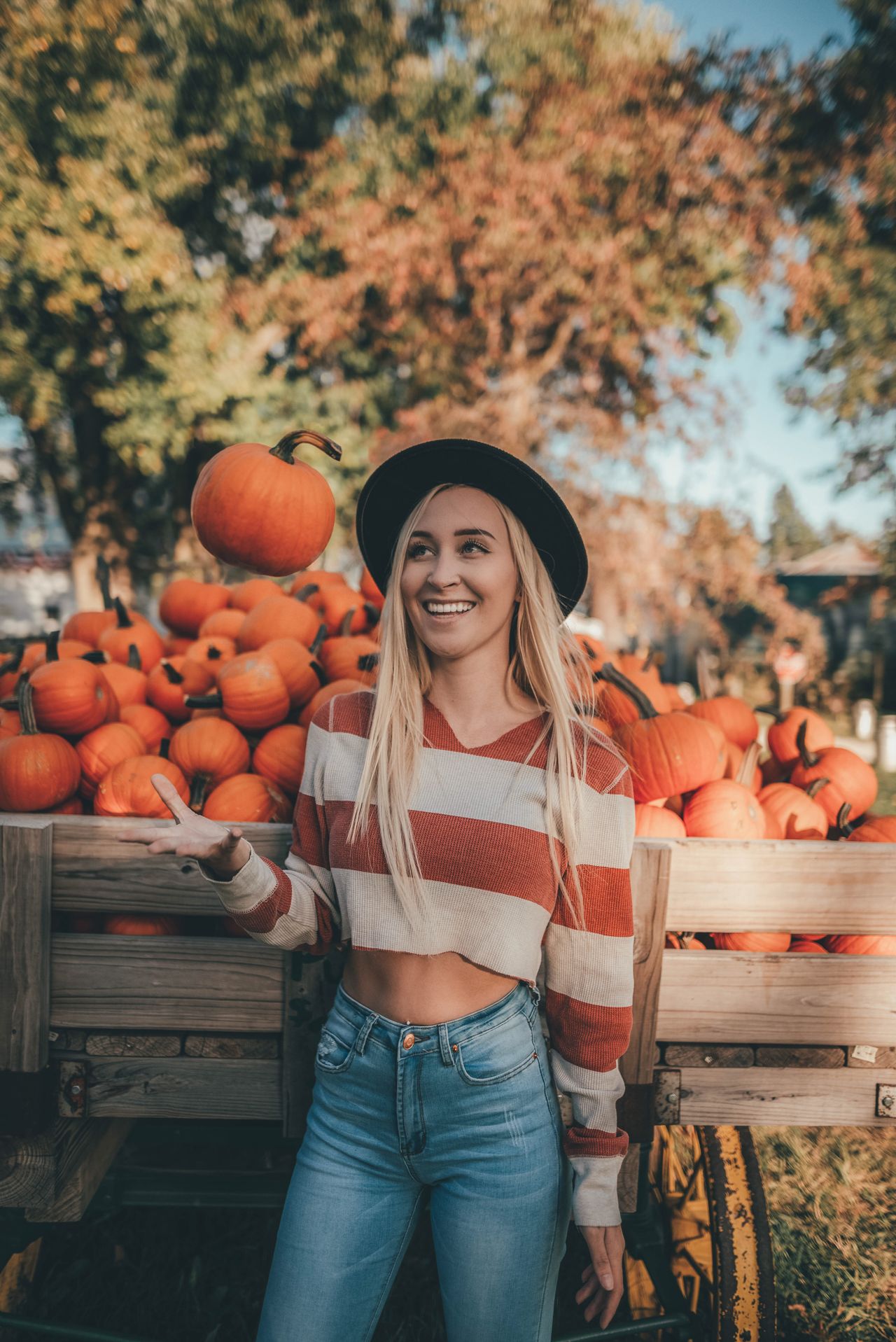 Woman in a striped sweater and felt hat playfully tossing a pumpkin in front of a wagon of pumpkins