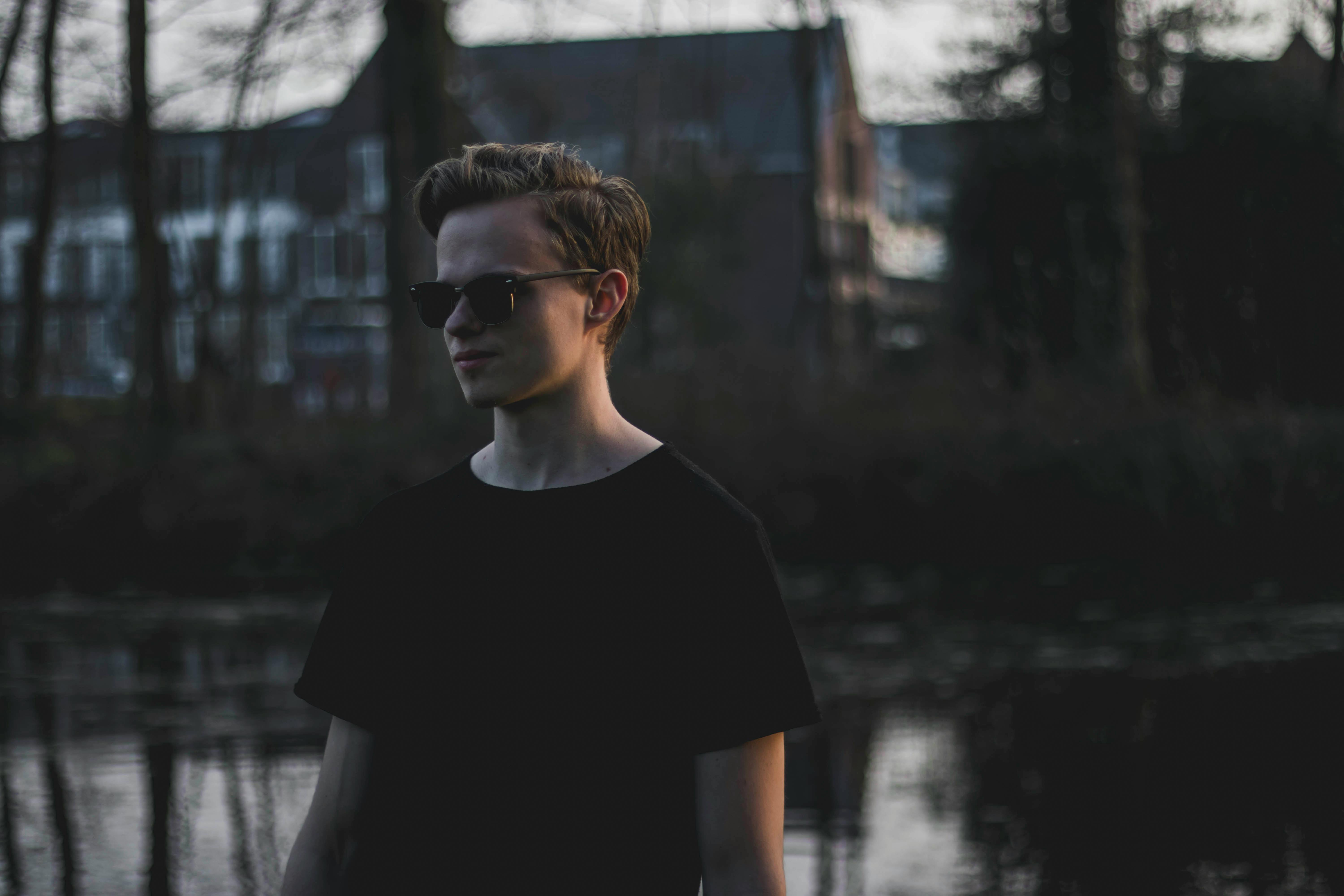 Portrait of a young man wearing sunglasses standing by a serene lake in a reflective pose.
