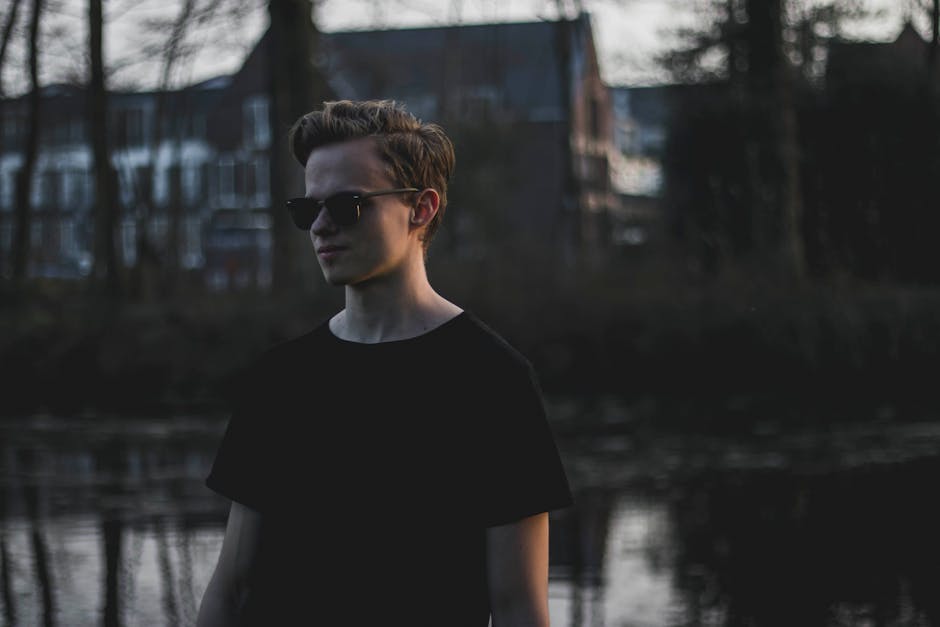 Portrait of a young man wearing sunglasses standing by a serene lake in a reflective pose.