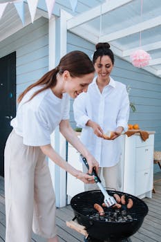 Two women enjoy grilling sausages outdoors during a cheerful summer BBQ party.