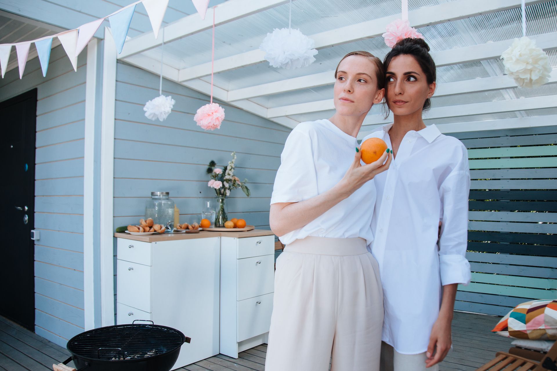 Two women in white outfits stand together in a decorated outdoor patio with oranges and bunting.