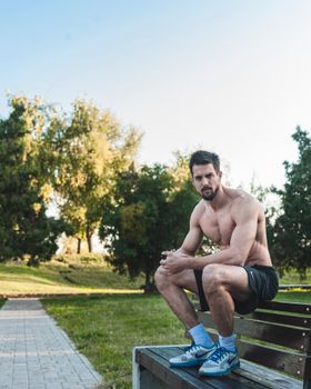 Fit adult man with a beard sitting shirtless on a bench in a sunny park.