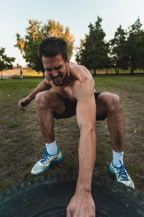 Free Fit man in athletic gear squatting outdoors performing a tire workout during daylight. Stock Photo