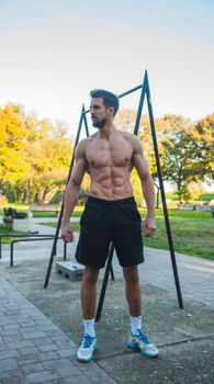 A shirtless muscular man training at an outdoor gym in the park on a sunny day.