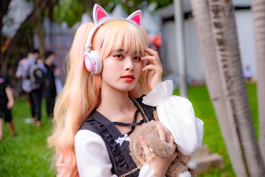 Young woman in cosplay attire with cat ear headphones posing outdoors holding a plush toy.