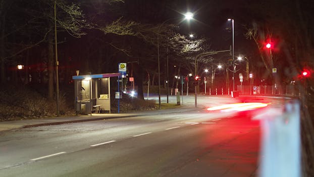 一条安静的夜间城市街道上，设有公交车站和路灯，过往汽车的车灯划出条条光带