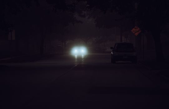 A car's headlights piercing the darkness on a quiet street at night, with a mysterious and moody atmosphere.