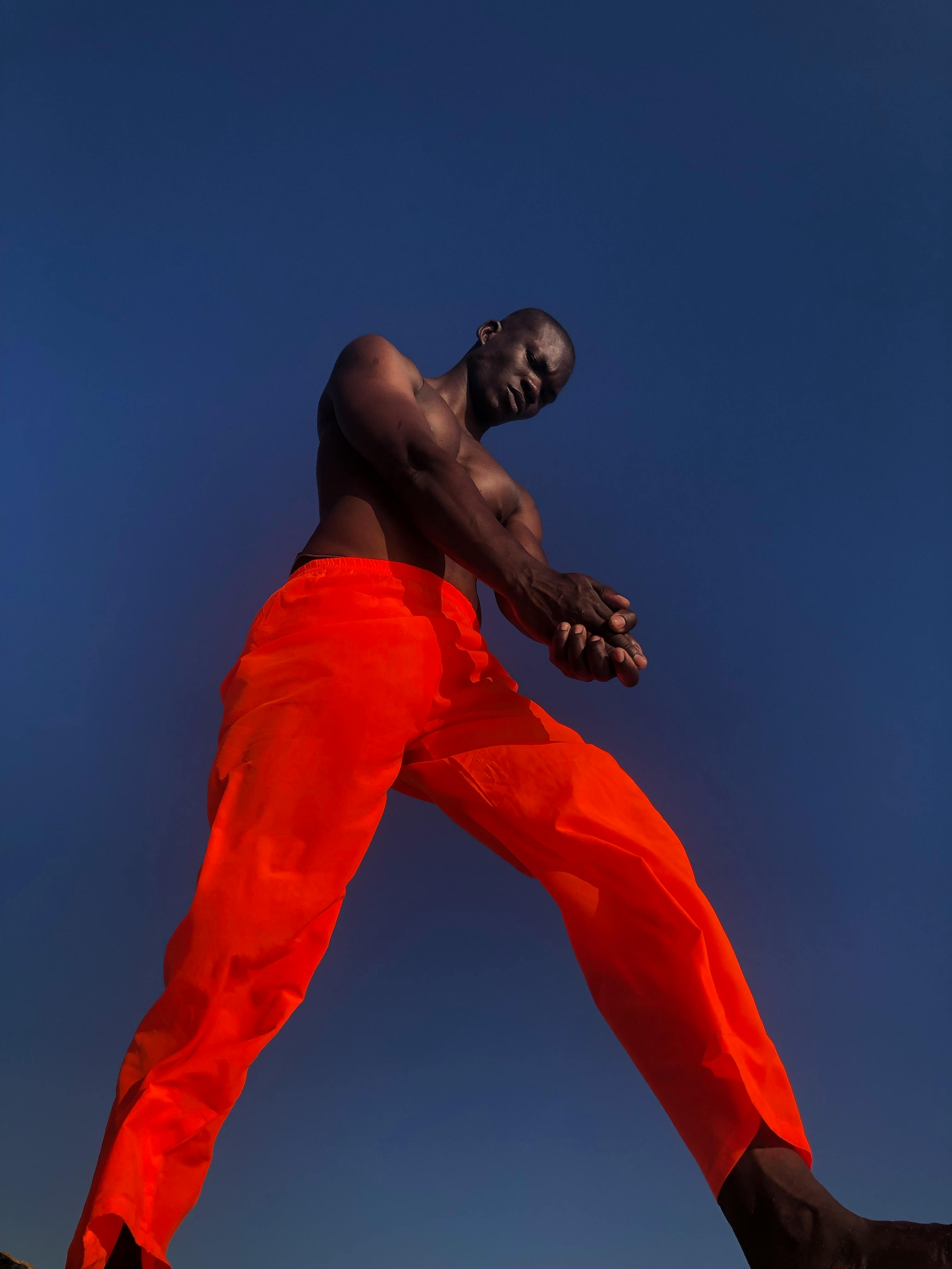A powerful perspective of a shirtless man in orange pants against a deep blue sky.