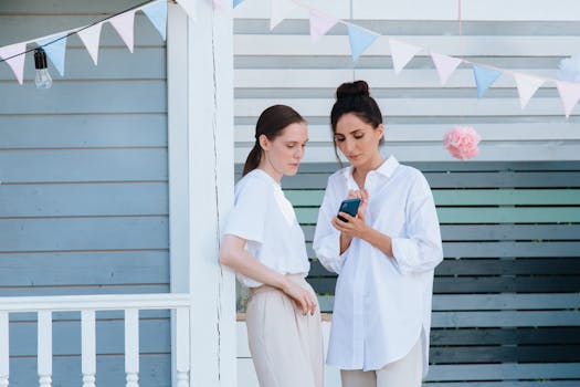 Two women in casual attire interacting with a cellphone outdoors under festive decorations.