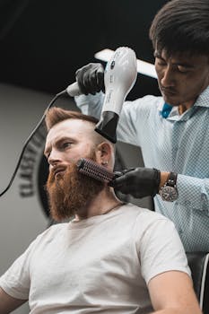 Barber using a hairdryer and brush to style a customer's beard in a modern barbershop.