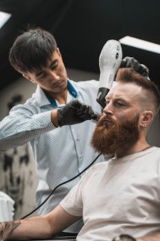 Barber styling a bearded man's hair with a blow dryer in a modern barbershop.