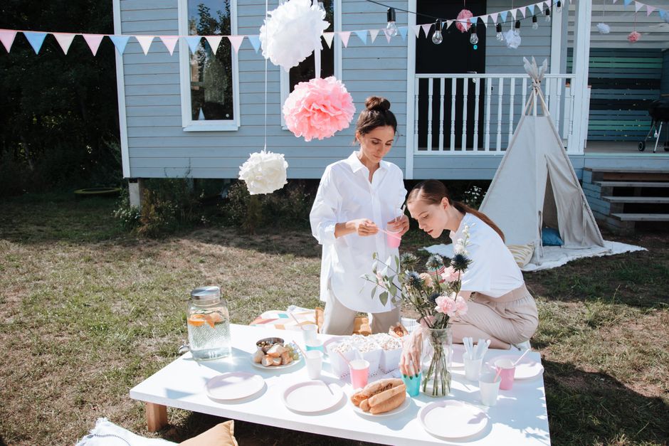 Two women setting up an outdoor summer party table with snacks and decorations.