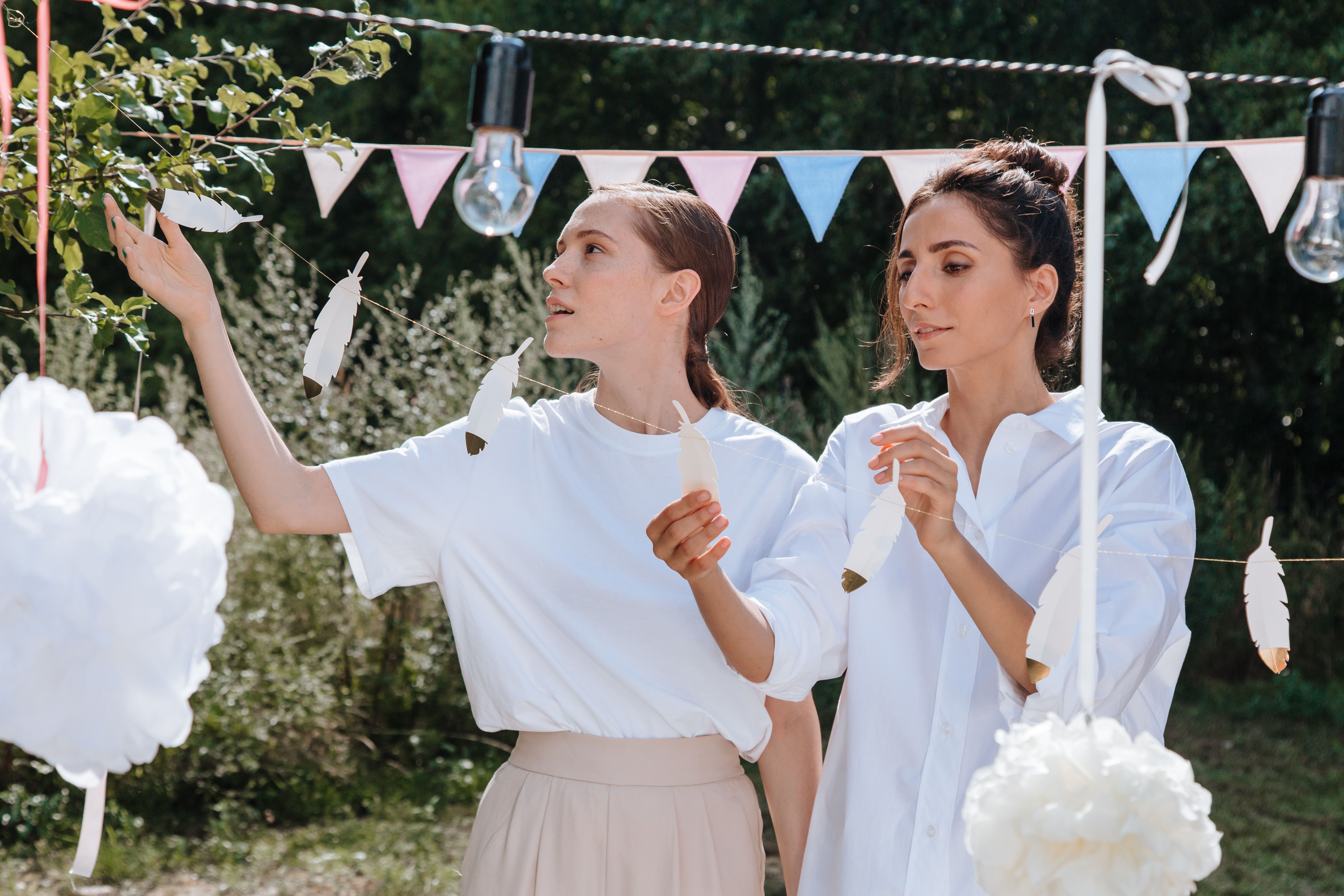 Two women decorating with feathers at an outdoor celebration. Summer vibes with festive banners.