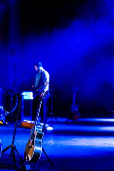 A vibrant blue-lit stage with an acoustic guitar and performer in soft focus.