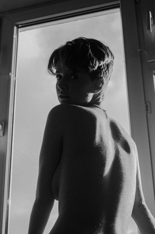 Free A monochrome portrait of a shirtless woman looking back through a window, highlighting artistic nudity. Stock Photo