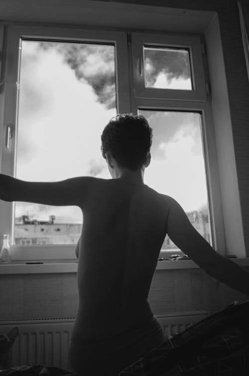 Grayscale Photo of a Shirtless Woman Standing Near a Window
