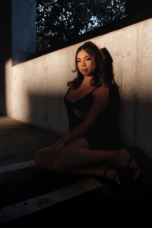 Free Asian woman in black dress posing in a shadowed urban environment, sitting against a wall. Stock Photo