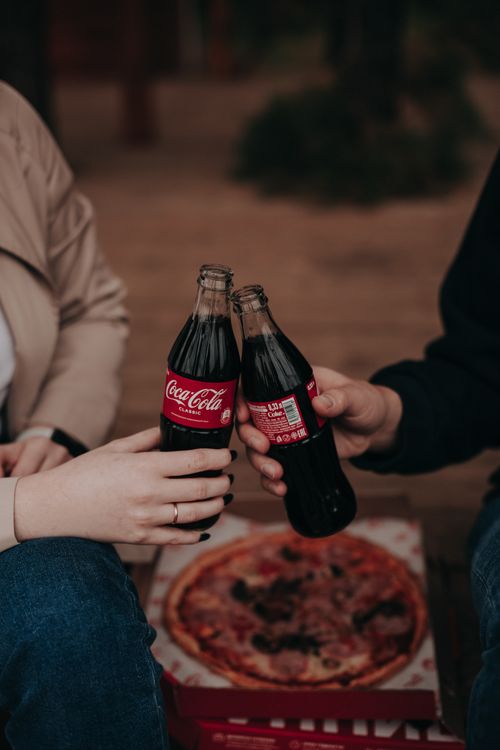 Free Two people toast with Coca-Cola bottles over a pizza. Warm, social setting. Stock Photo