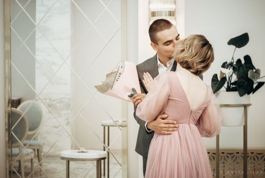 A couple shares a romantic kiss indoors, holding a bouquet of flowers.