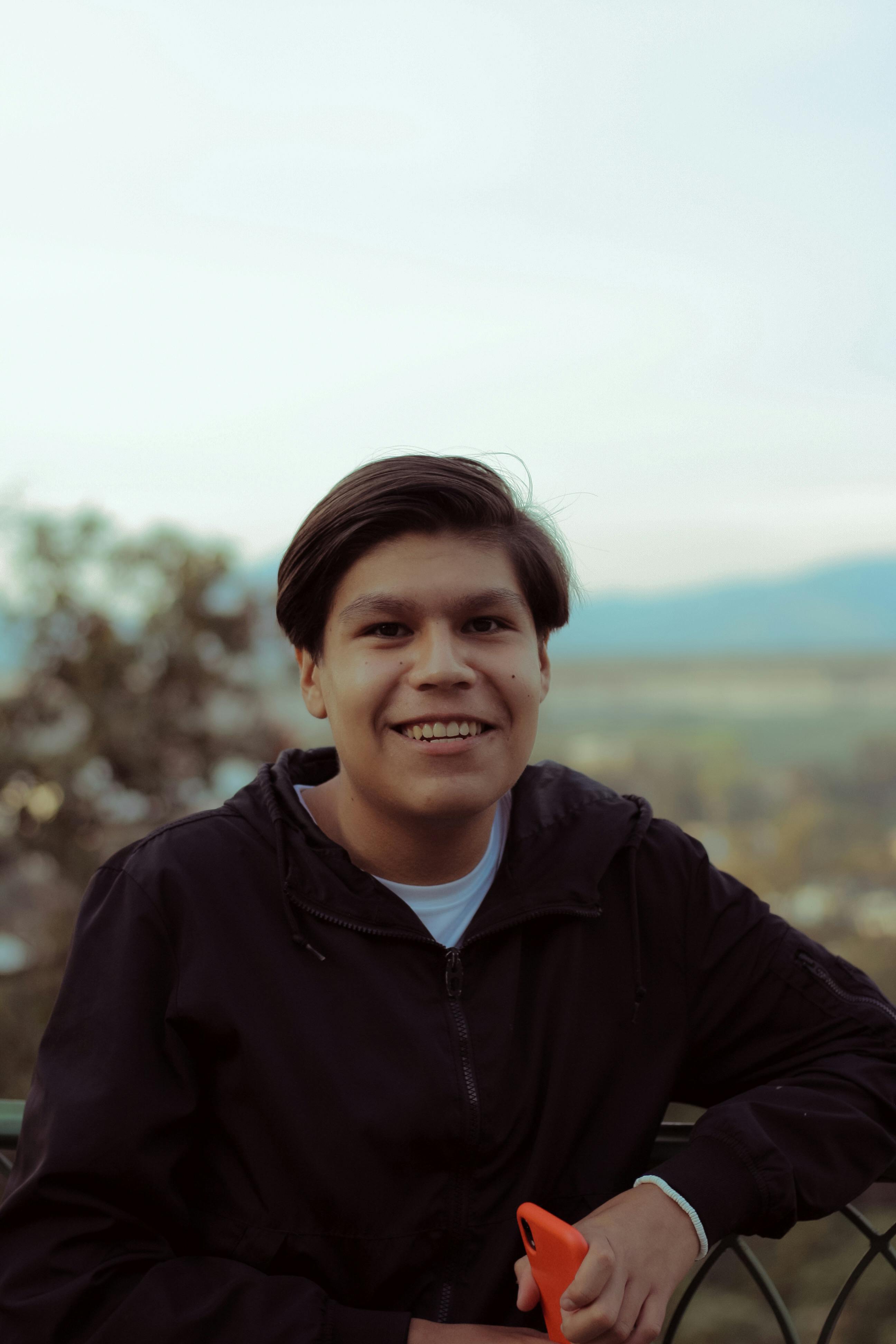 Smiling young man in black jacket holding smartphone outdoors during the day.