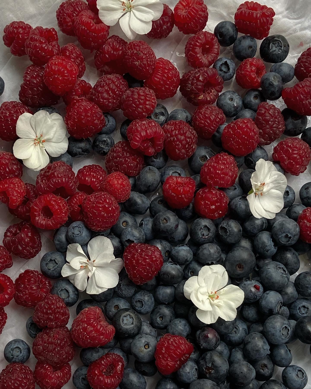 A vibrant flat lay of fresh blueberries, raspberries, and white flowers on a light background.