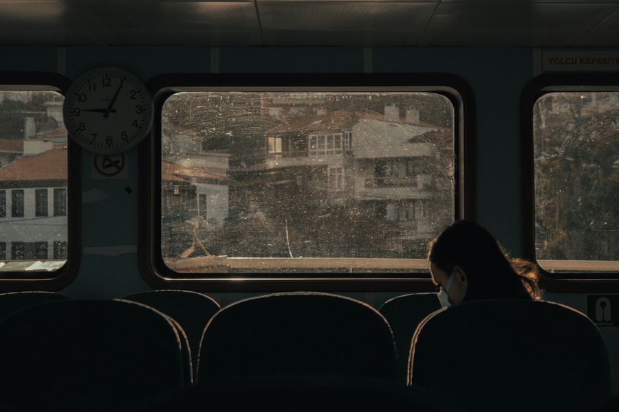 A pensive woman in a mask sits inside a train, gazing at a cityscape through dirt-speckled windows.