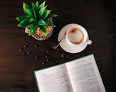 一张舒适的咖啡休息场景的顶视图，木桌上摆着一本打开的书、一个咖啡杯和一些多汁蔬菜