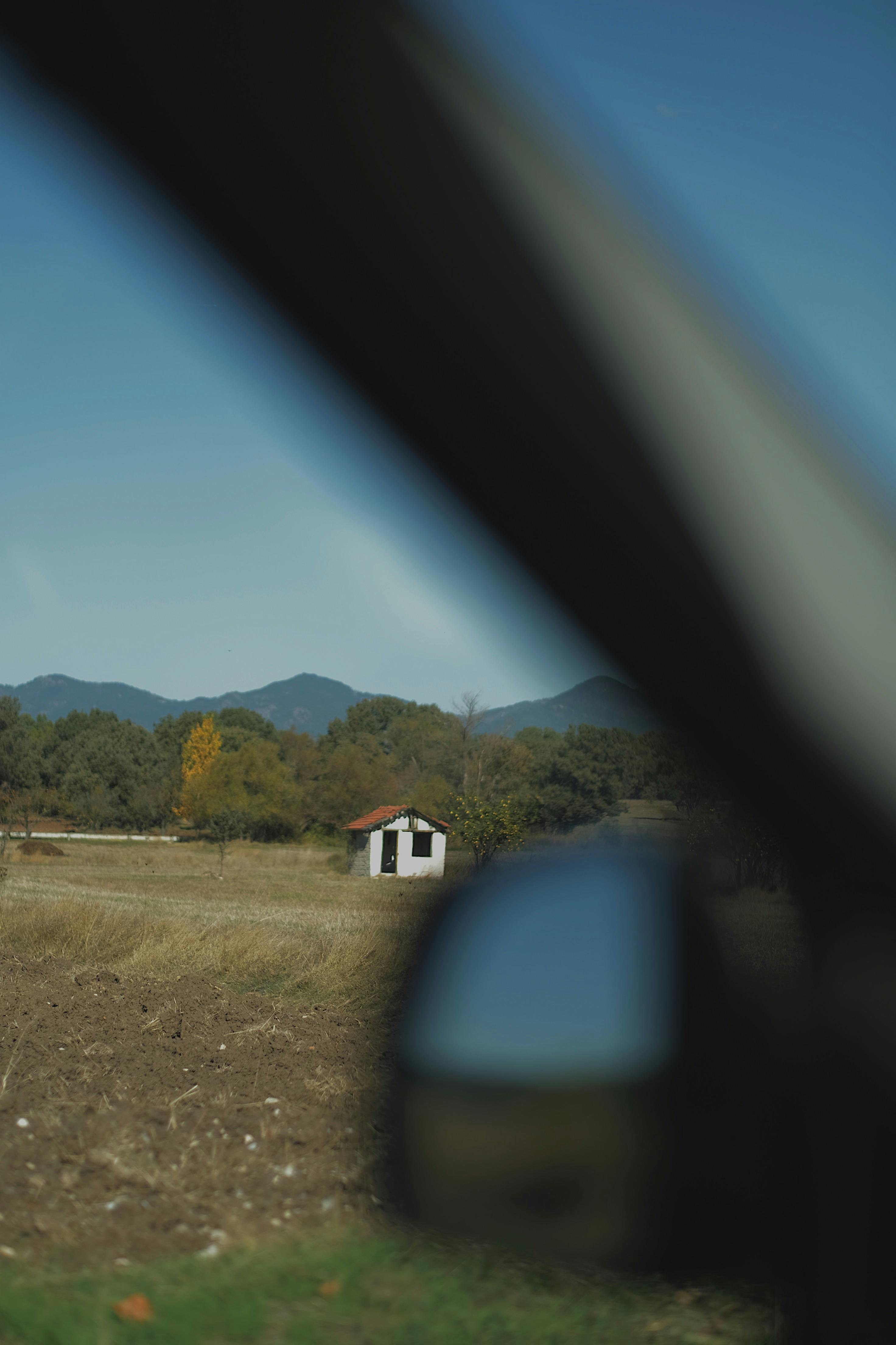 A picturesque rural scene captured through a vehicle window, featuring a distant house and mountain backdrop.