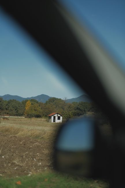 A picturesque rural scene captured through a vehicle window, featuring a distant house and mountain backdrop.