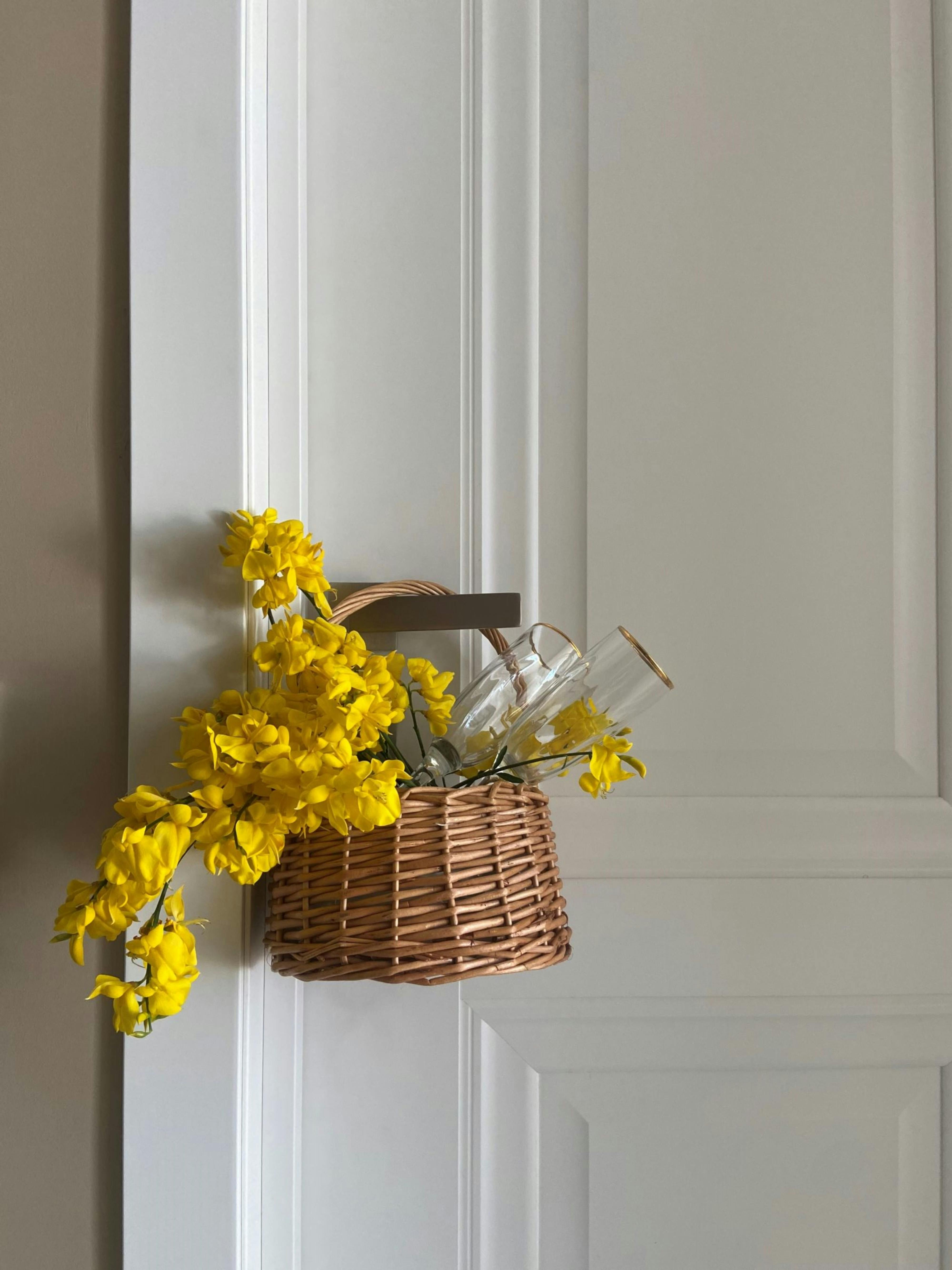 A wicker basket with yellow flowers and wine glasses hangs gracefully on a white door.