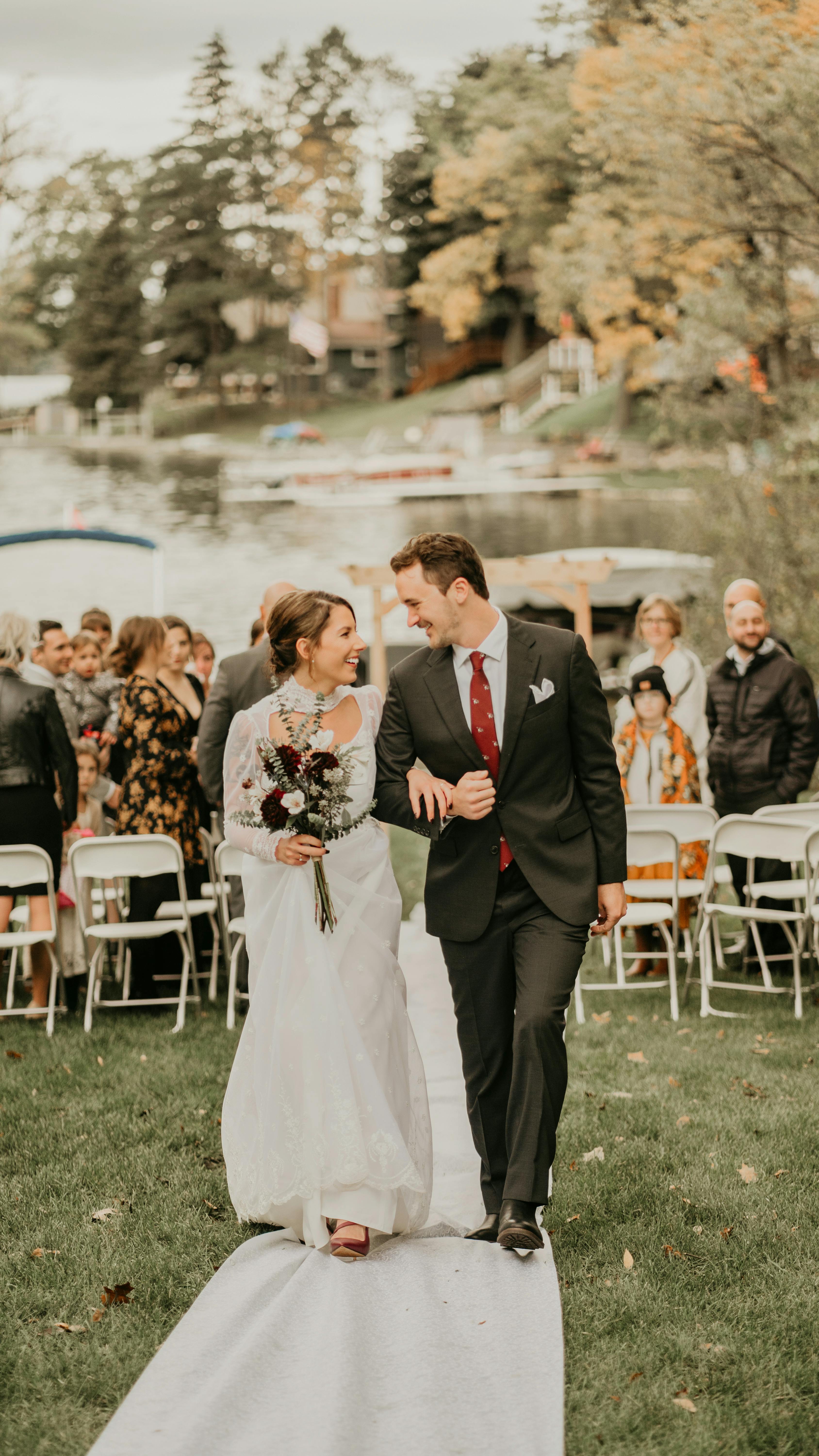 Bride and groom walking down the aisle outdoors by a lake, smiling at each other.