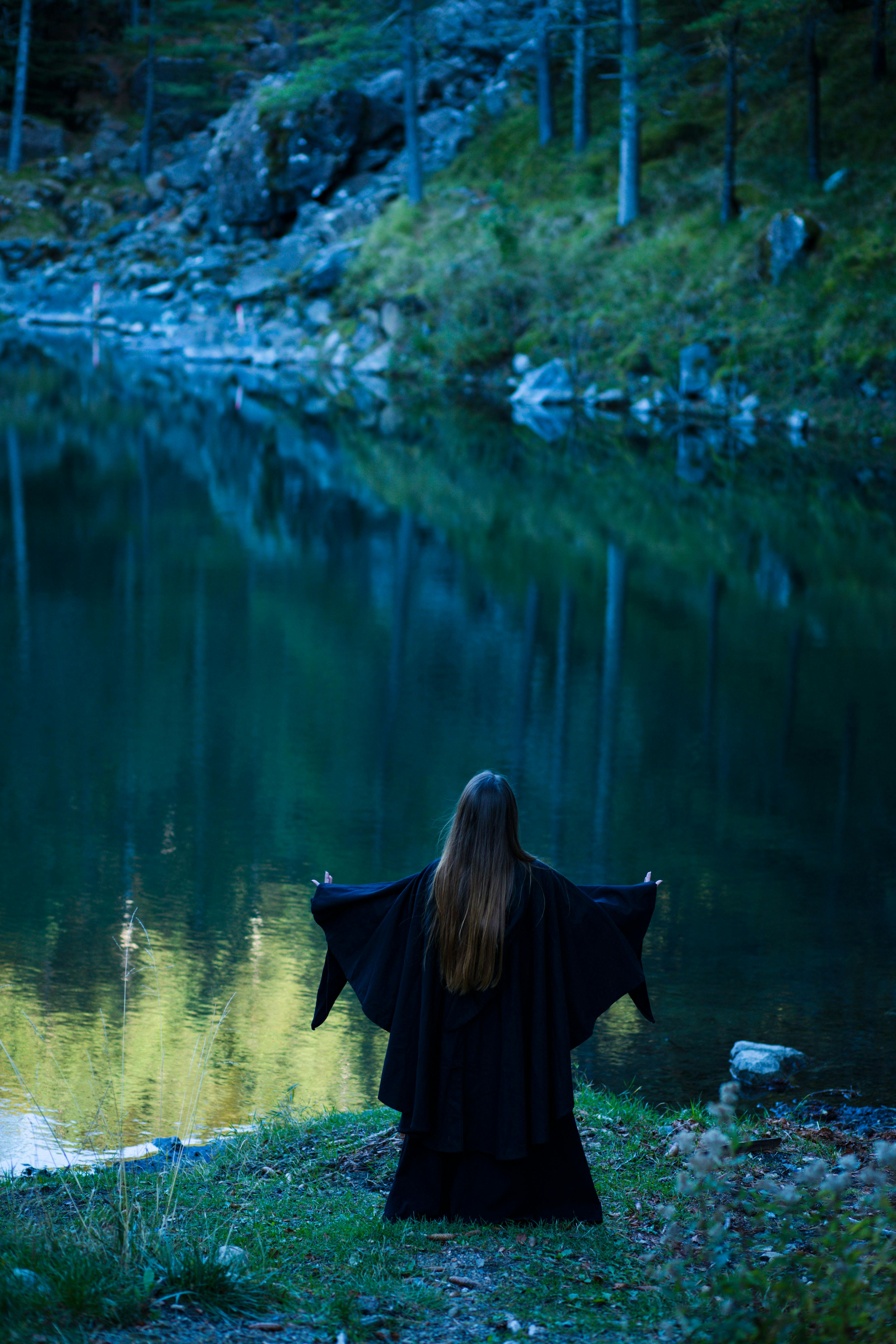 A mysterious woman in a black cloak stands by a tranquil forest lake.
