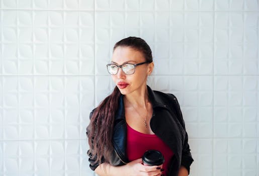 Portrait of a fashionable woman in glasses holding a coffee cup against a white tiled background.