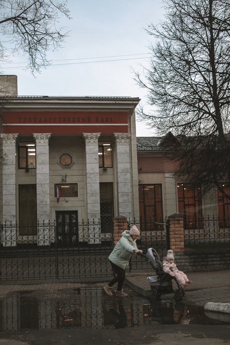 A Woman In Winter Clothing Pushing The Stroller While Walking On The Sidewalk Of The Street