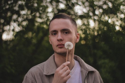 A young man with a dandelion against a blurred green background, fashionably styled.
