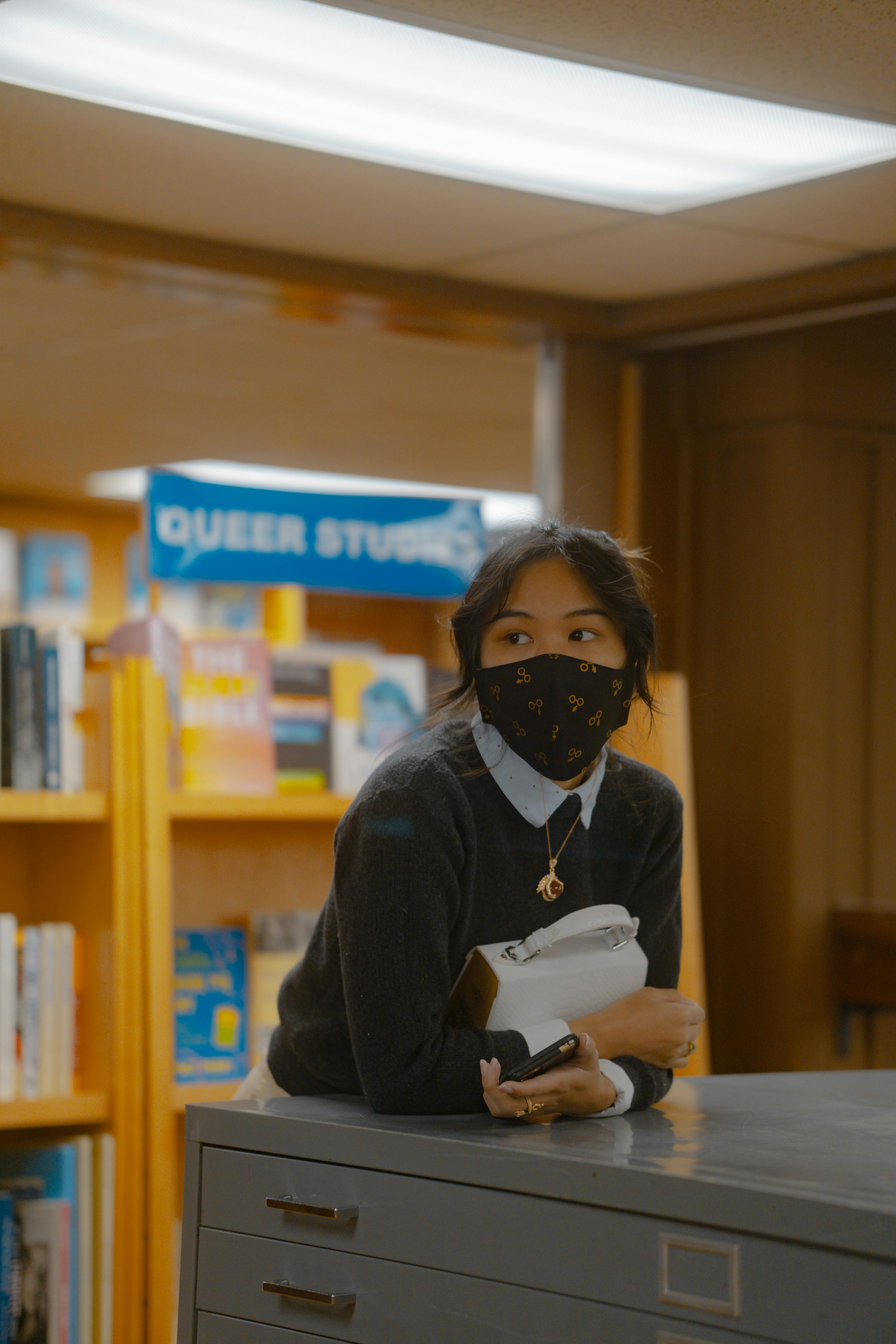 Free Asian woman wearing a mask leans on a drawer in a library's queer studies section. Stock Photo