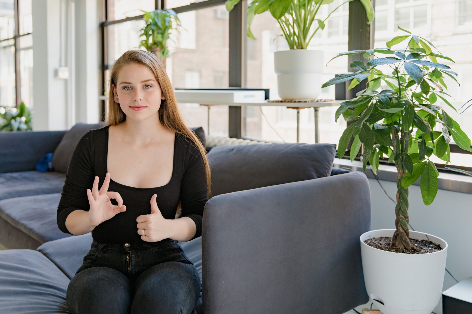 Young woman sitting indoors with plants, gesturing OK sign, bright setting.