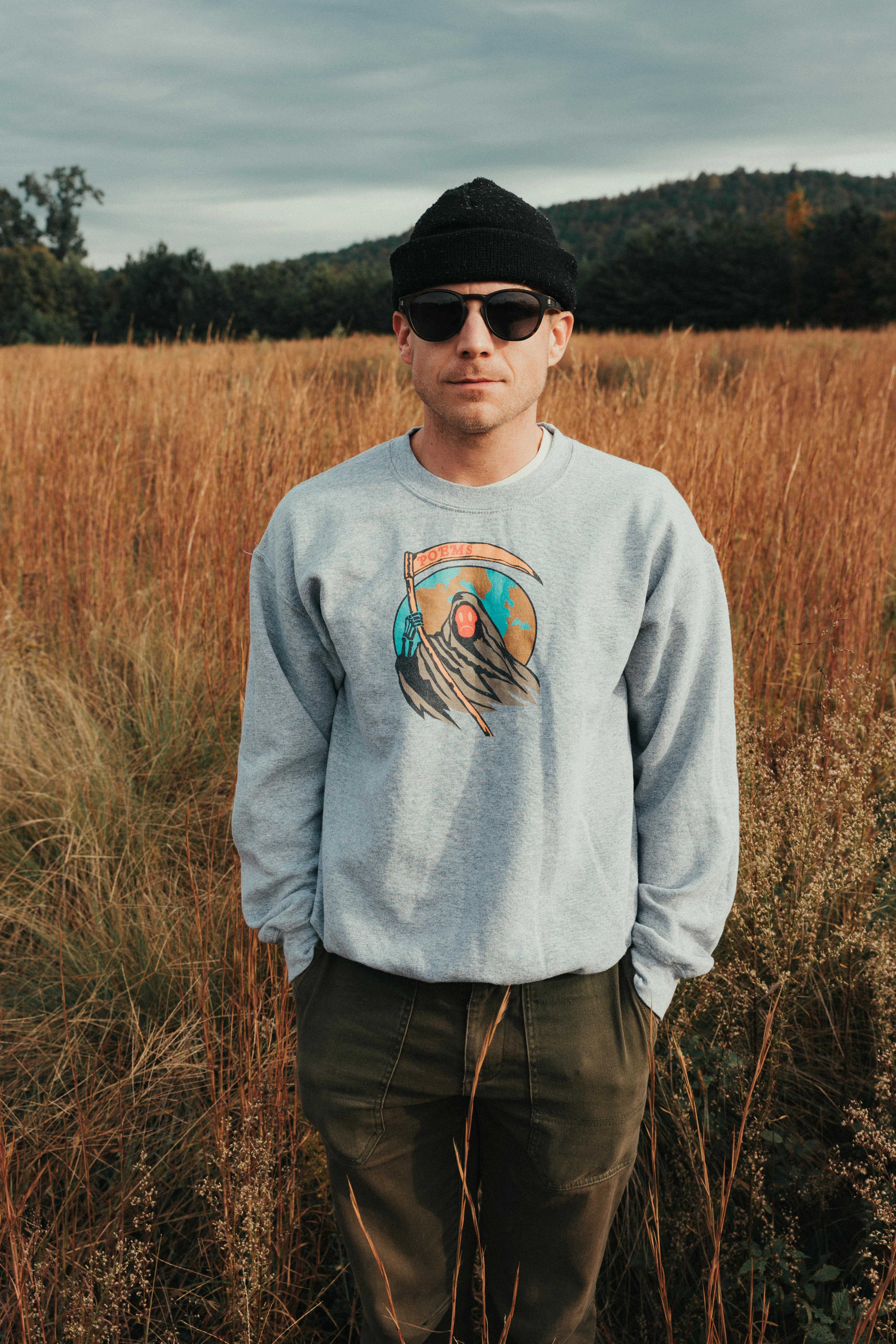 Man standing in a field wearing sunglasses, hat, and graphic sweatshirt, exuding a relaxed vibe.