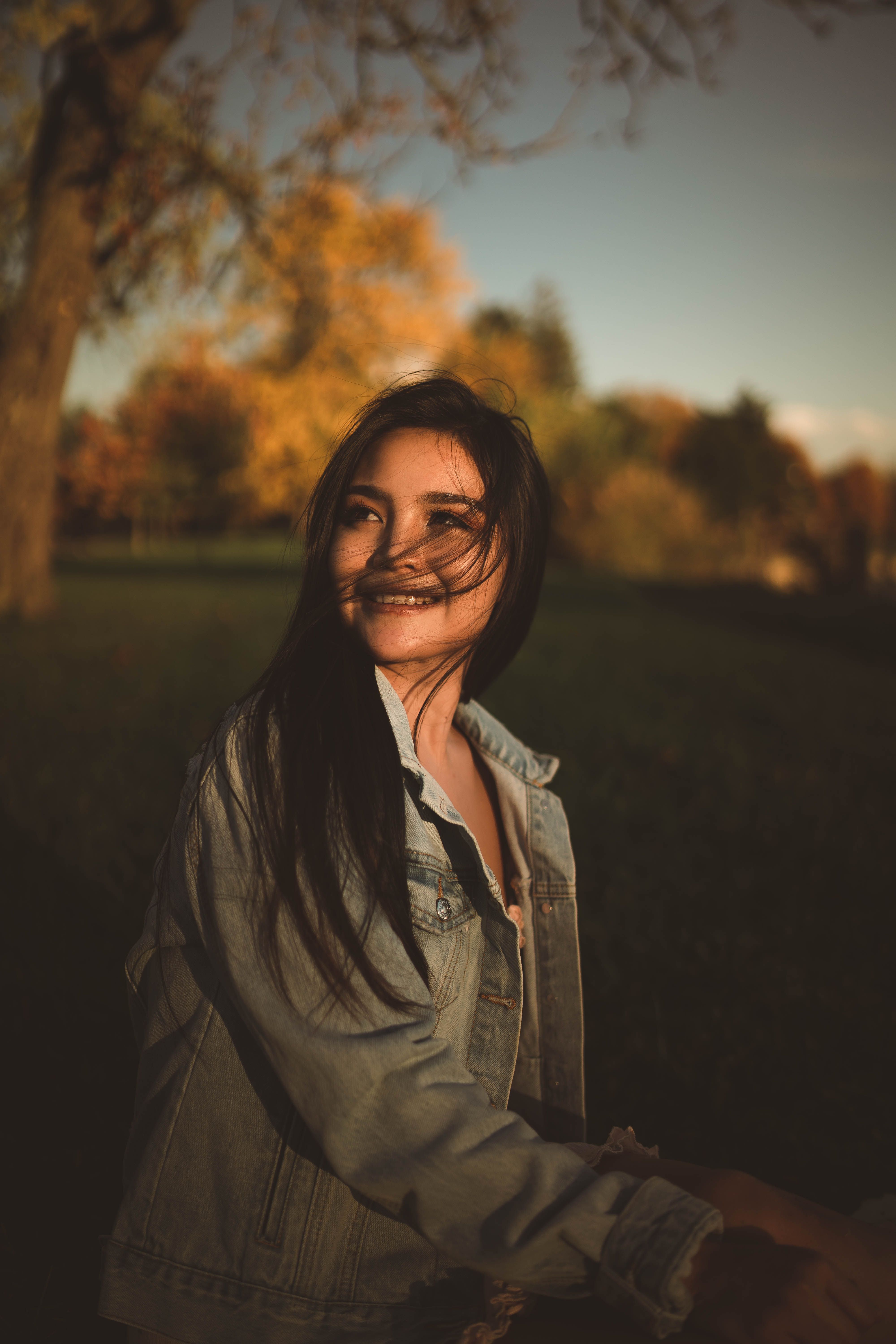 Smiling woman in denim jacket enjoying a sunset in a park during autumn.
