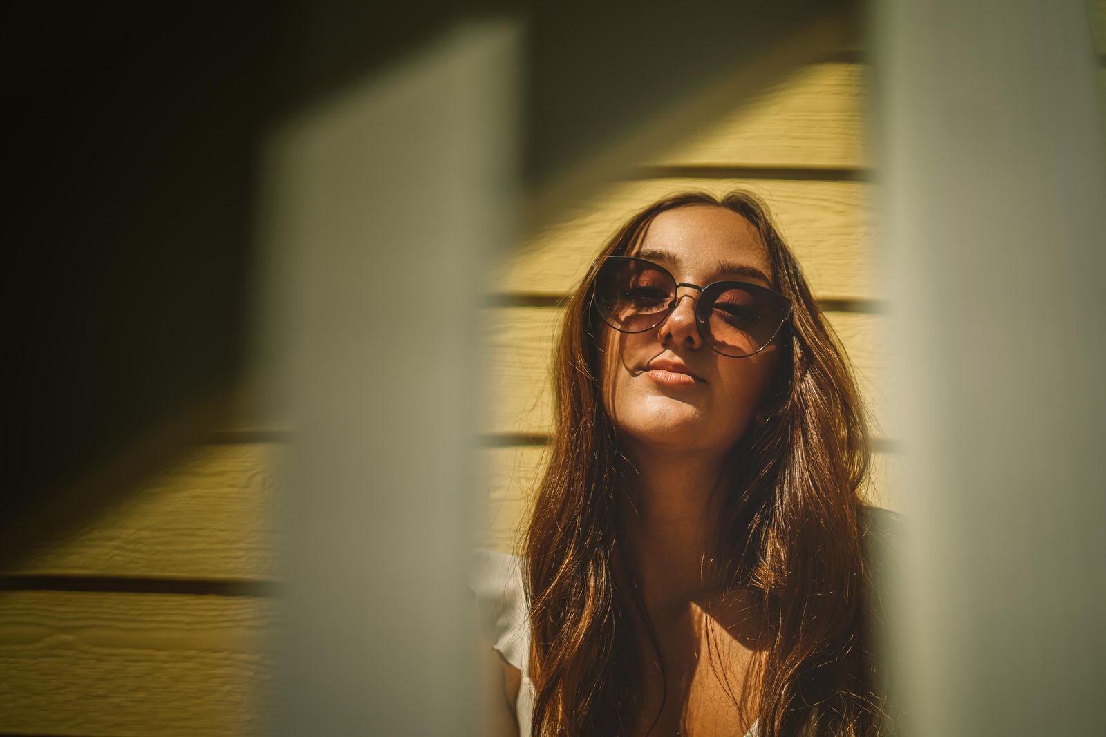 Woman with long brown hair and round sunglasses tilting her face toward warm sunlight beside a yellow wall
