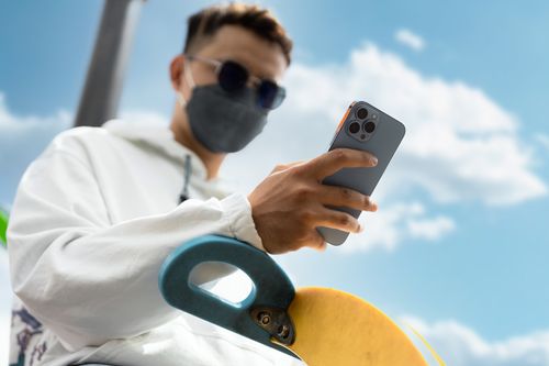 Free A young man wearing a face mask uses his cellphone outdoors with a skateboard in the foreground. Stock Photo
