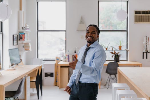 Smiling businessman in a shirt and tie giving a thumbs up in a bright, modern office setting.