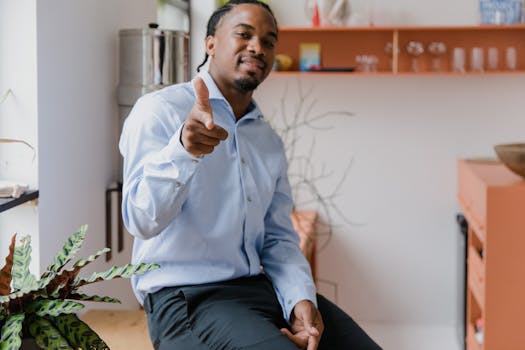 Confident businessman in a blue shirt gesturing at the camera while sitting in a modern office setting.
