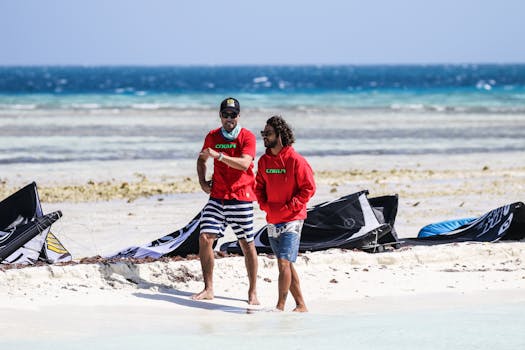 Two men in casual attire walking along a sunny beach with kite surfing equipment in view.