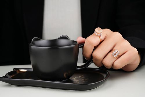Free Close-up of a hand adorned with rings, holding a stylish black cat-themed cup on a tray. Sophisticated jewelry display. Stock Photo