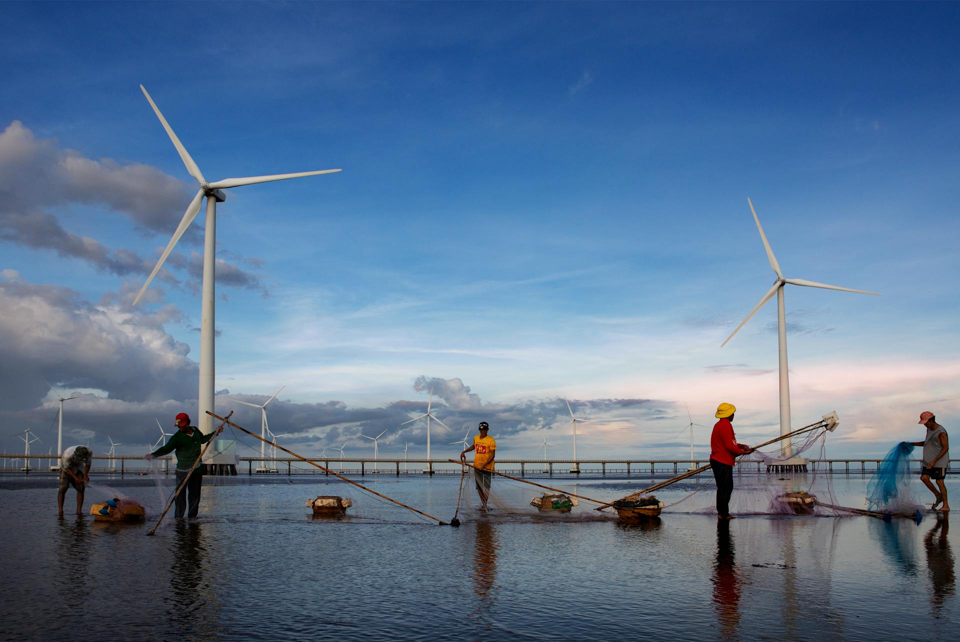 A group of fishermen cast nets near wind turbines set against a vibrant sunset sky, showcasing sustainable energy.