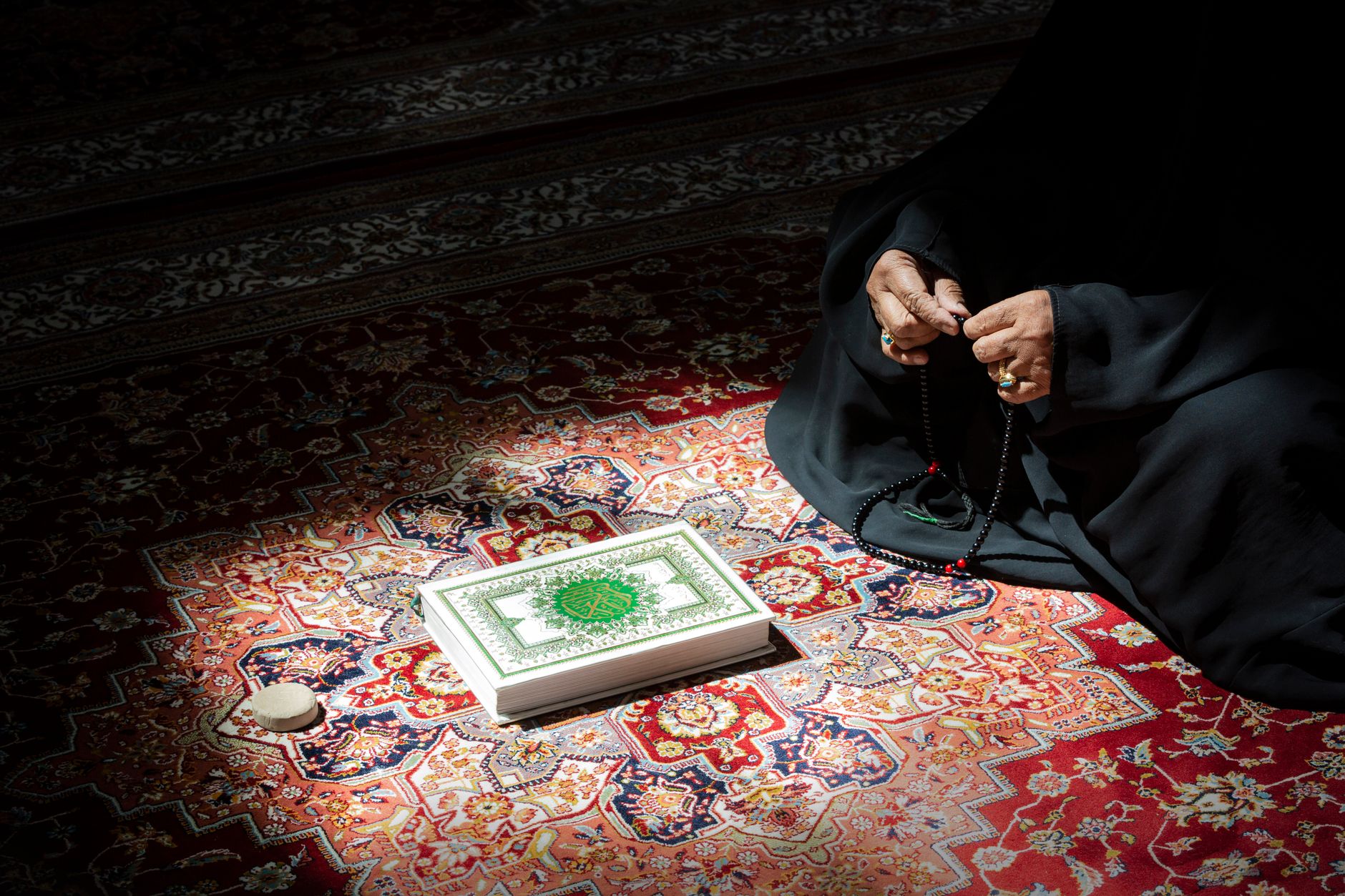 A woman in serene prayer holding beads beside an ornate Quran on a colorful rug.