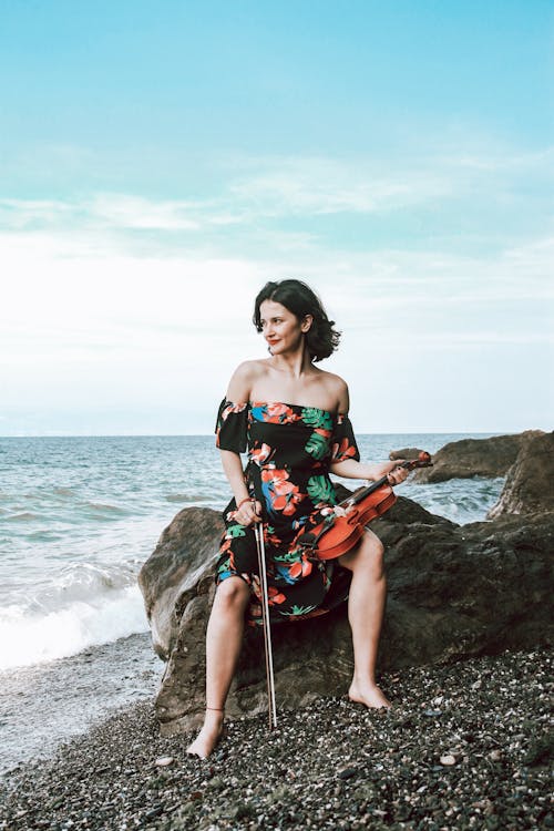 Free Woman in Black Dress Sitting on Rock While Holding a Violin Stock Photo
