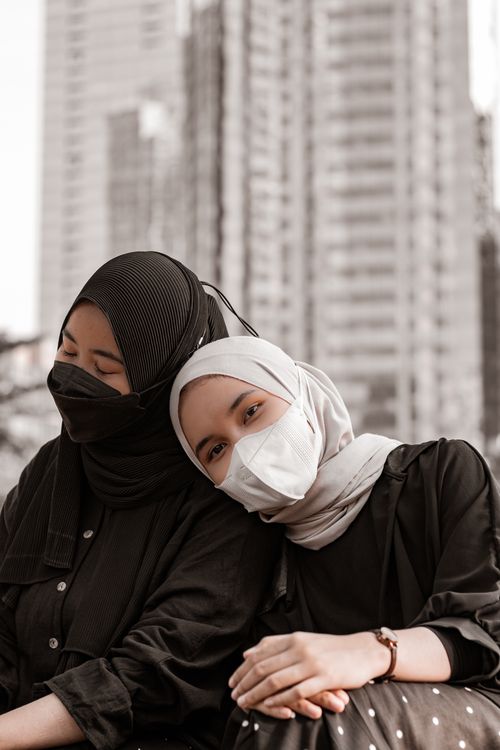 Free Two women wearing hijabs and face masks sitting closely together in an urban setting. Stock Photo